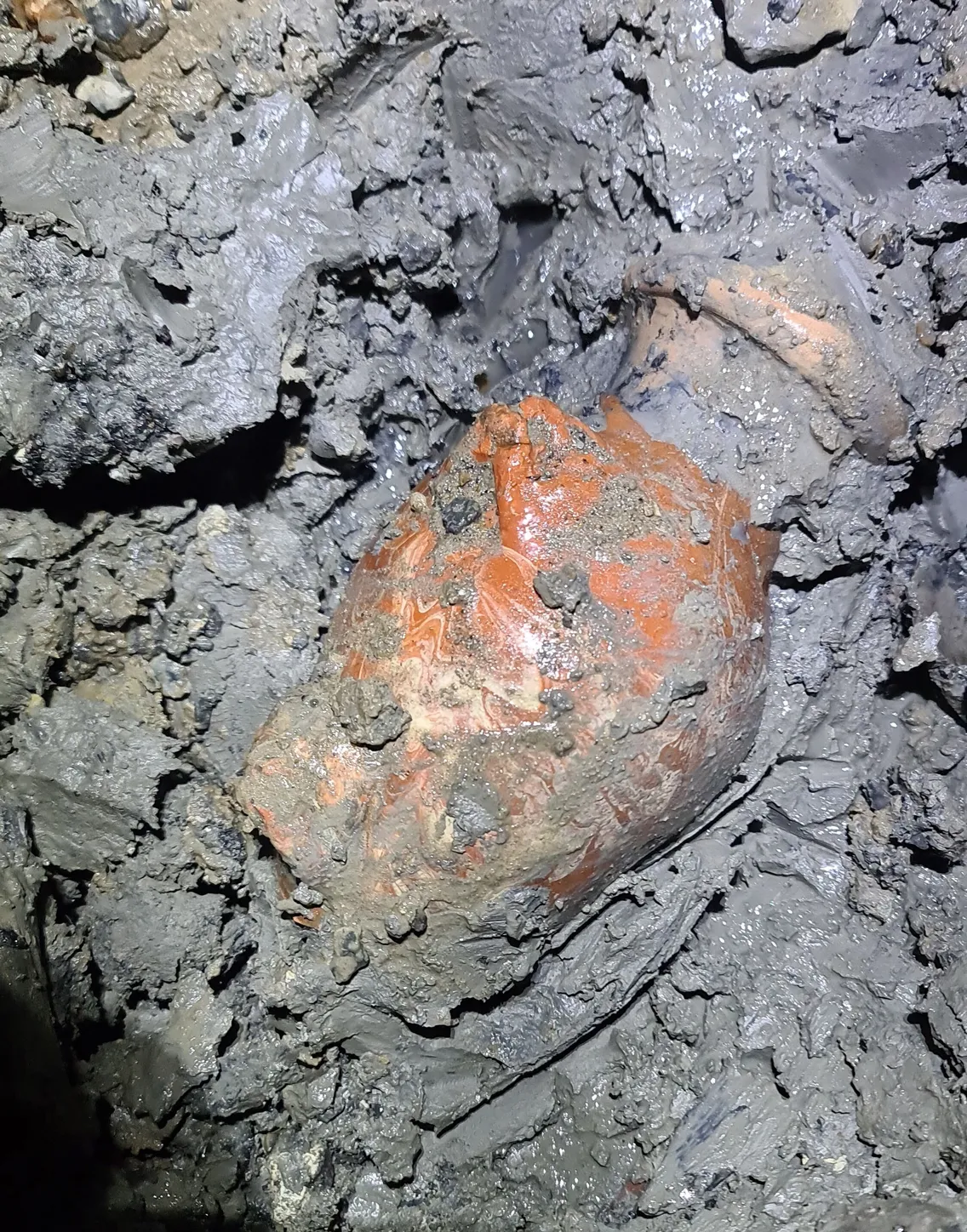 A partially buried clay pot is exposed in wet, muddy soil.