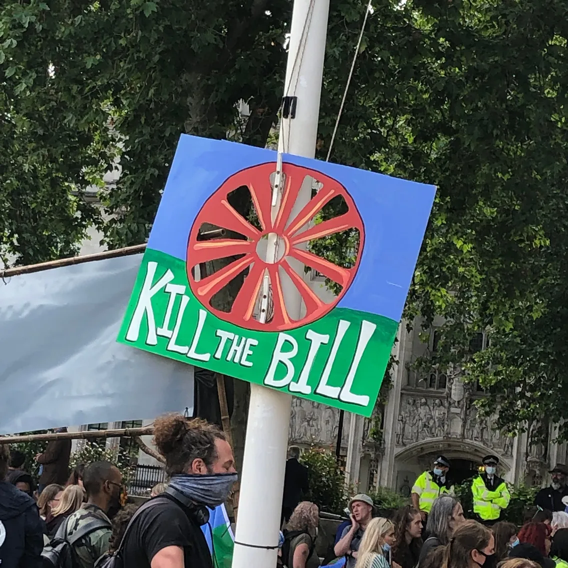 A protest sign reading "Kill the Bill" with a red wheel symbol is held up in front of a crowd, with police officers and trees in the background.