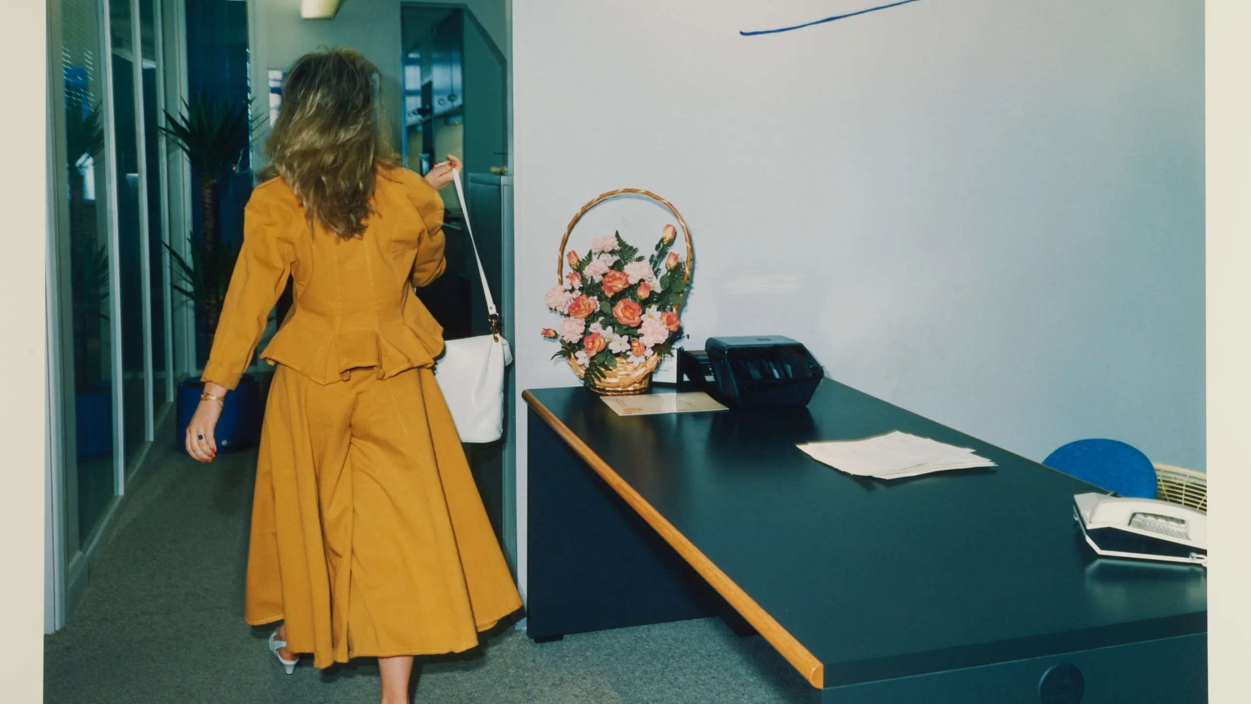 Woman in a mustard coloured suit walking away in an office environment (description, not title).

From the series ‘Work Stations’, co-commissioned by the Museum of London and Camerawork in 1987, documenting the new office culture of the 1980s and the emerging technologies that rapidly transformed the nature of the working day. This was the first body of photographic work looking at office life in Britain (diverging from the more typical documentary photography of the working classes) and it captures the mundanity, competition, rivalry, stress and gender imbalances of this new culture from a woman’s point of view. The images are accompanied by captions which are an integral part of the work. 