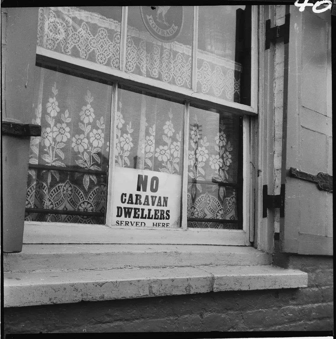 Black and white photo of a window with a "no caravan dwellers served here" sign on the sill, lace curtains visible behind the glass.