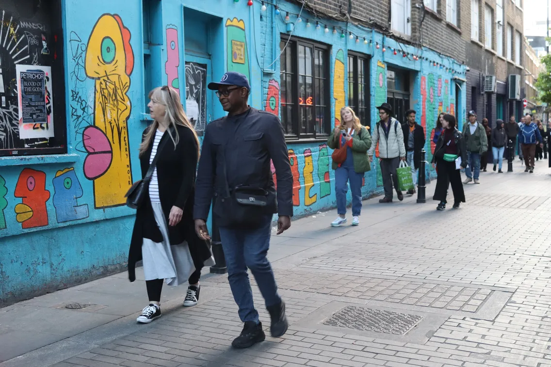 People walking along a street with a blue building covered in colorful graffiti art in the background.