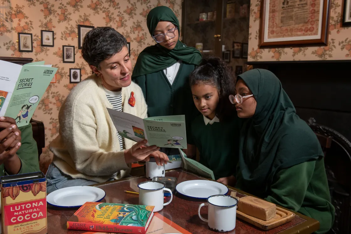 A woman and four girls, all wearing green uniforms, sit around a table with plates and books, reading together in a warmly lit room with floral wallpaper.
