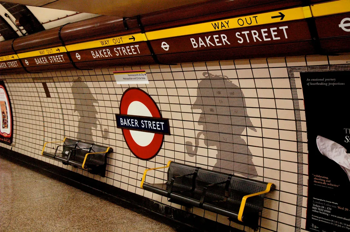 Baker Street Underground station platform with Sherlock Holmes silhouette art on tiled walls, benches below, and way out signs above.