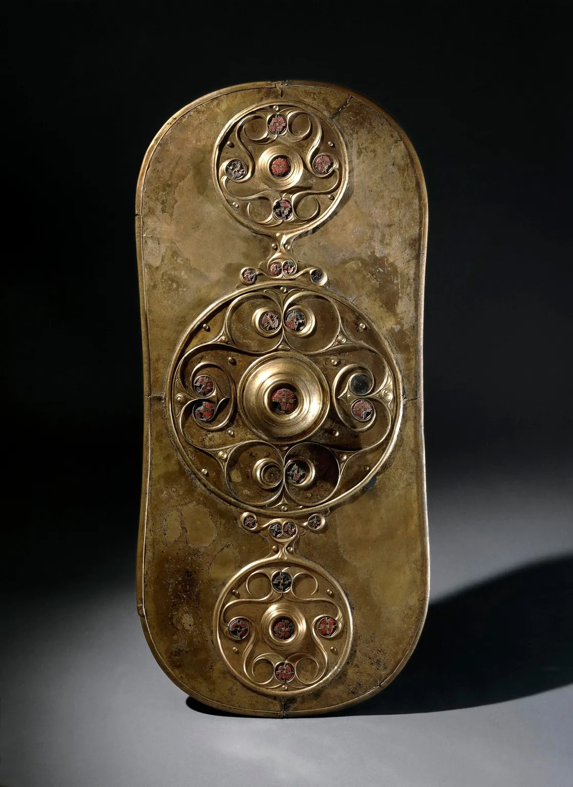 Bronze shield with three ornate circular designs and decorative red inlays, displayed against a dark background.