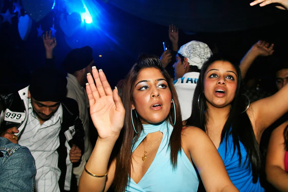 A group of young adults dance with their hands raised at a crowded indoor party, illuminated by blue lights.