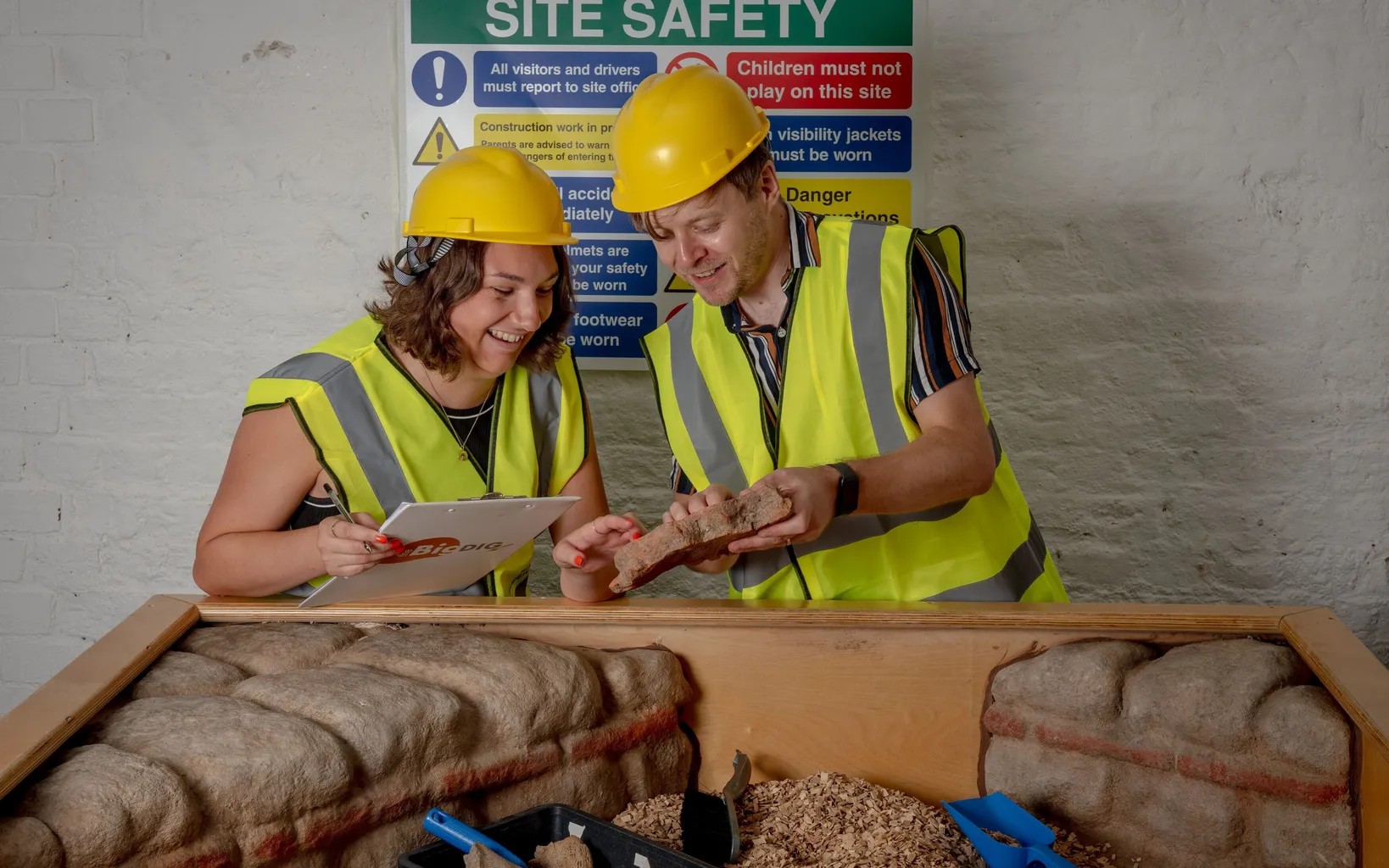 Two people in safety gear examine objects and take notes at an indoor excavation display, with a site safety sign visible in the background.