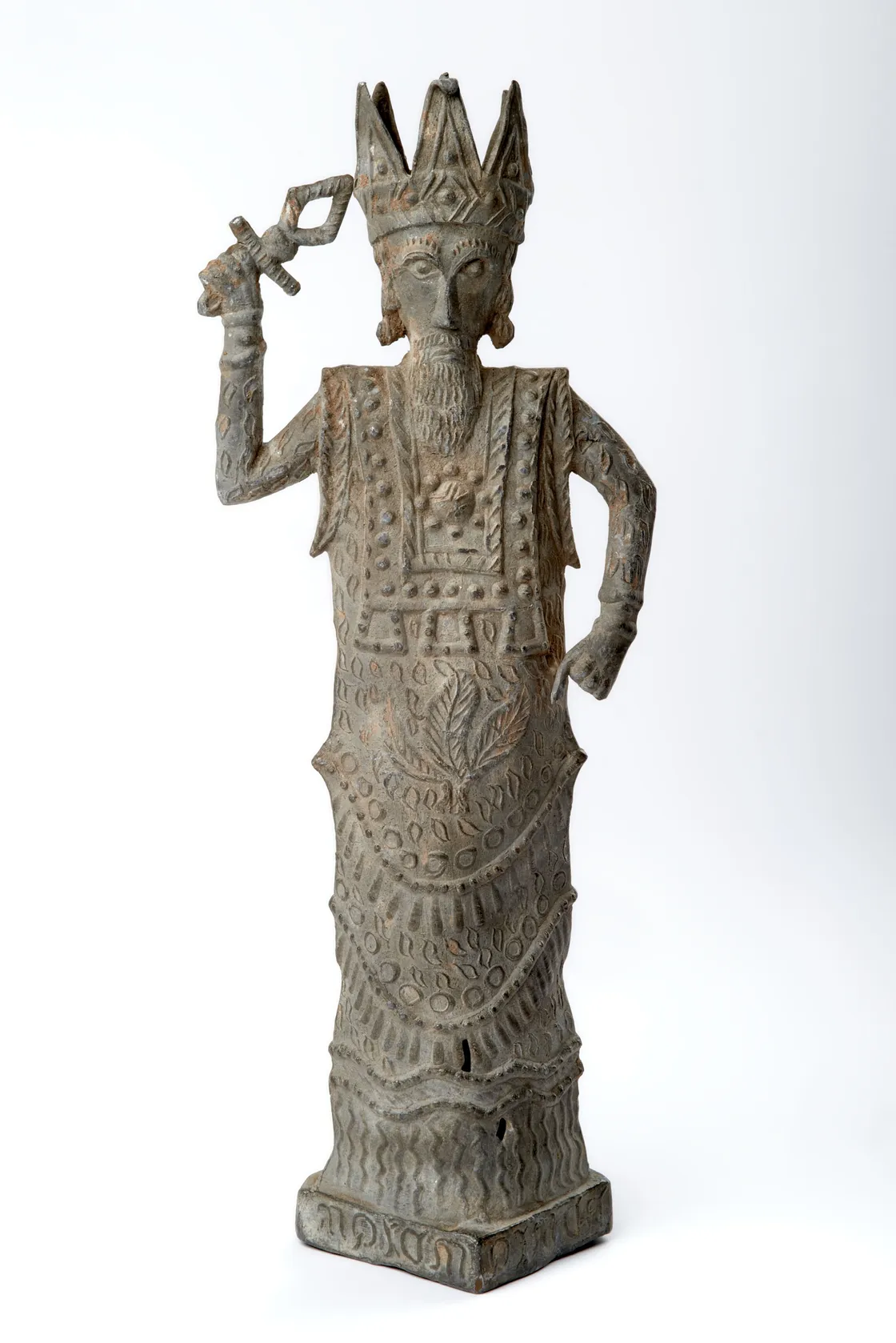 A metal statue of a bearded figure wearing a crown and ornate robe, holding an object in one hand, with the other hand resting on its hip.