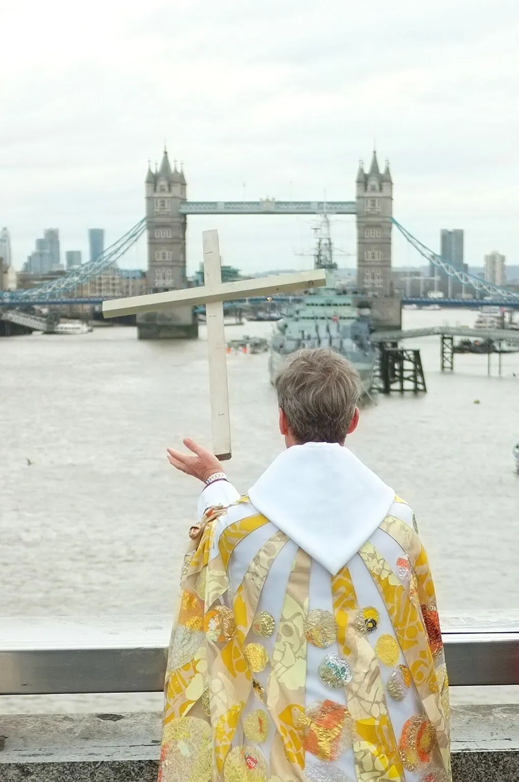 A person in ornate religious robes holds up a cross while facing the River Thames and Tower Bridge in London.