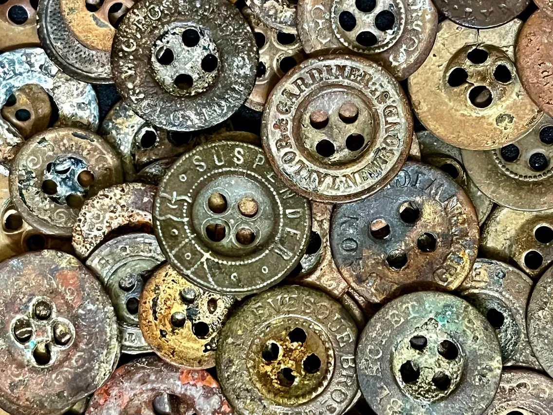 A close-up view of a pile of old, corroded metal buttons, some with engraved text and various levels of wear and patina.
