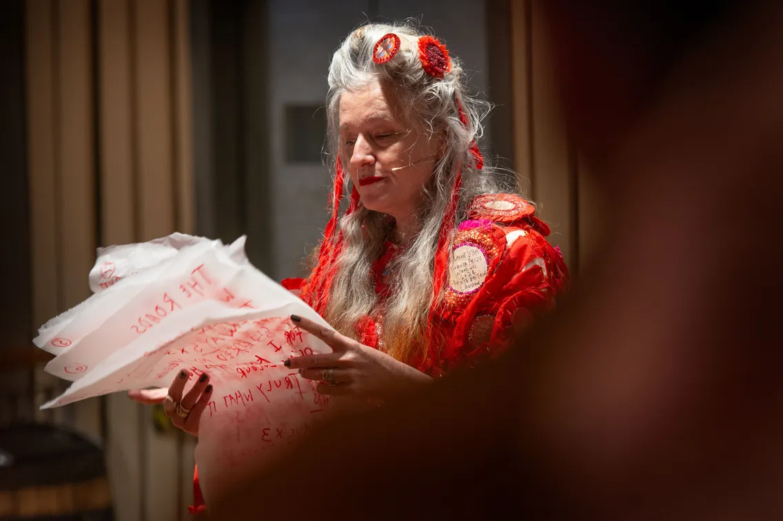 A person with long gray hair wearing a red outfit reads from large sheets of white paper covered in handwritten text.