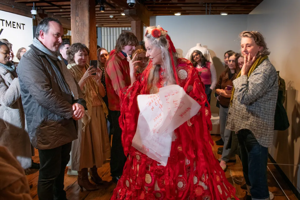 A woman in a red dress adorned with notes stands in the center of a group of people at an indoor event, while attendees look on and take photos.