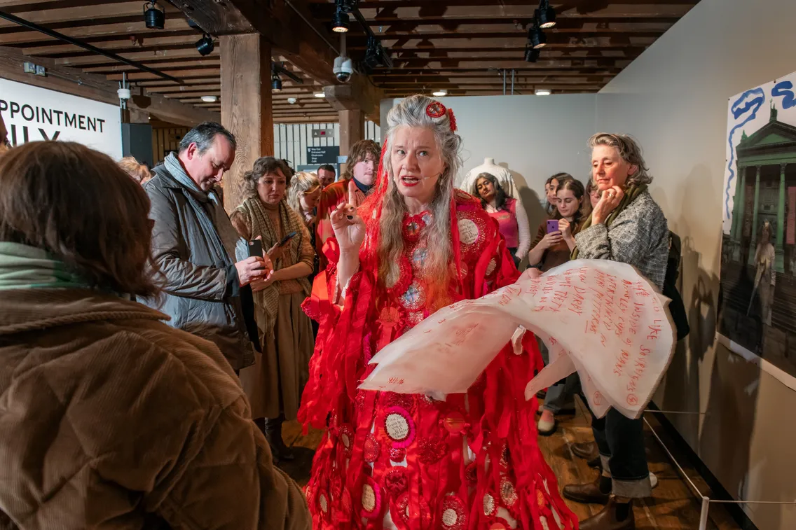A woman in a red costume with white writing stands in a gallery space, speaking to a group of people who are gathered around her.