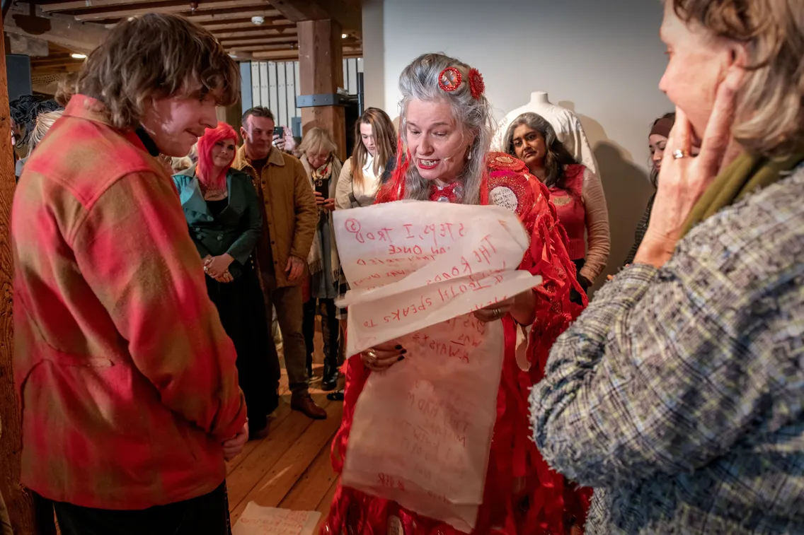 A woman in a red dress reads from large handwritten sheets to a small group of people indoors. Others listen attentively in the background.