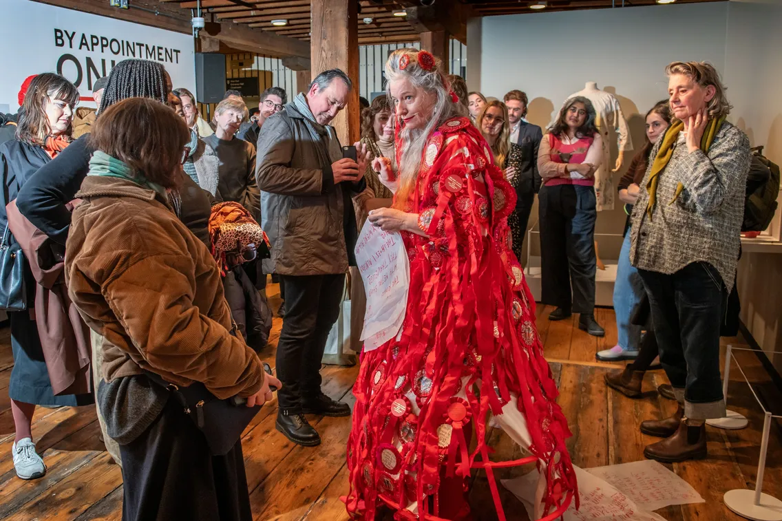 A woman in a red, elaborate costume stands in the center of a gallery, surrounded by a group of people observing her and reading papers attached to her outfit.