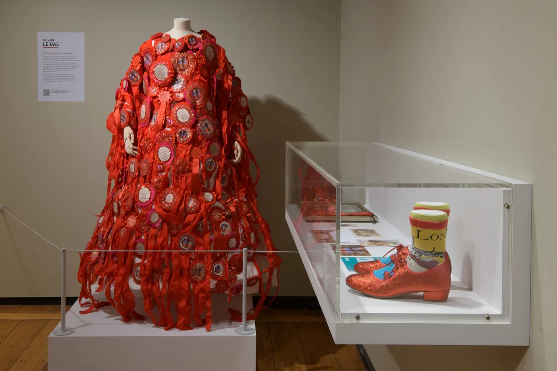 A mannequin displays a red, ribbon-covered dress with circular patches next to a glass case containing glittery red shoes and colorful socks.