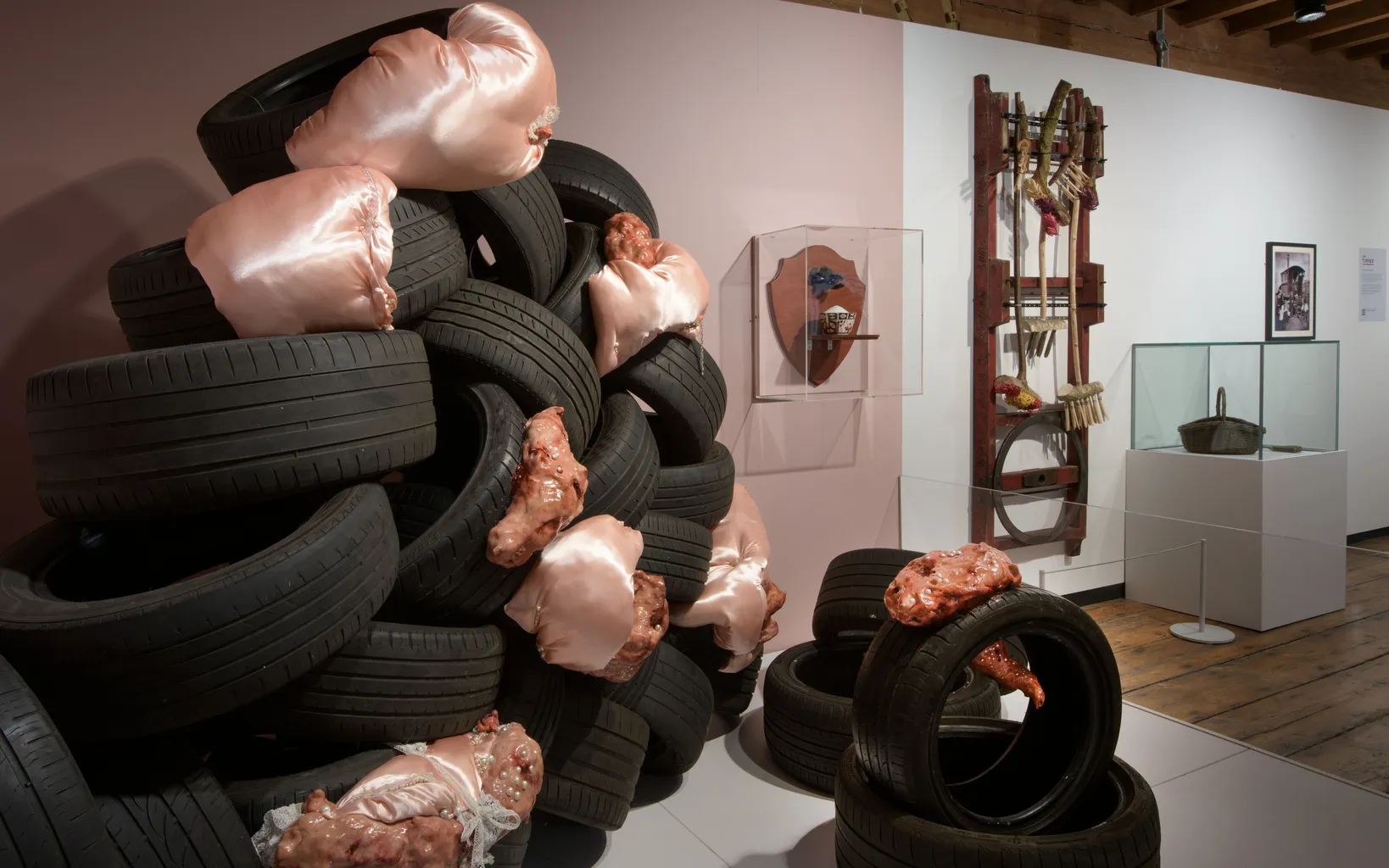 A stack of black tires interspersed with pink, flesh-like sculptures is displayed in an art gallery alongside tools and objects mounted on white walls.