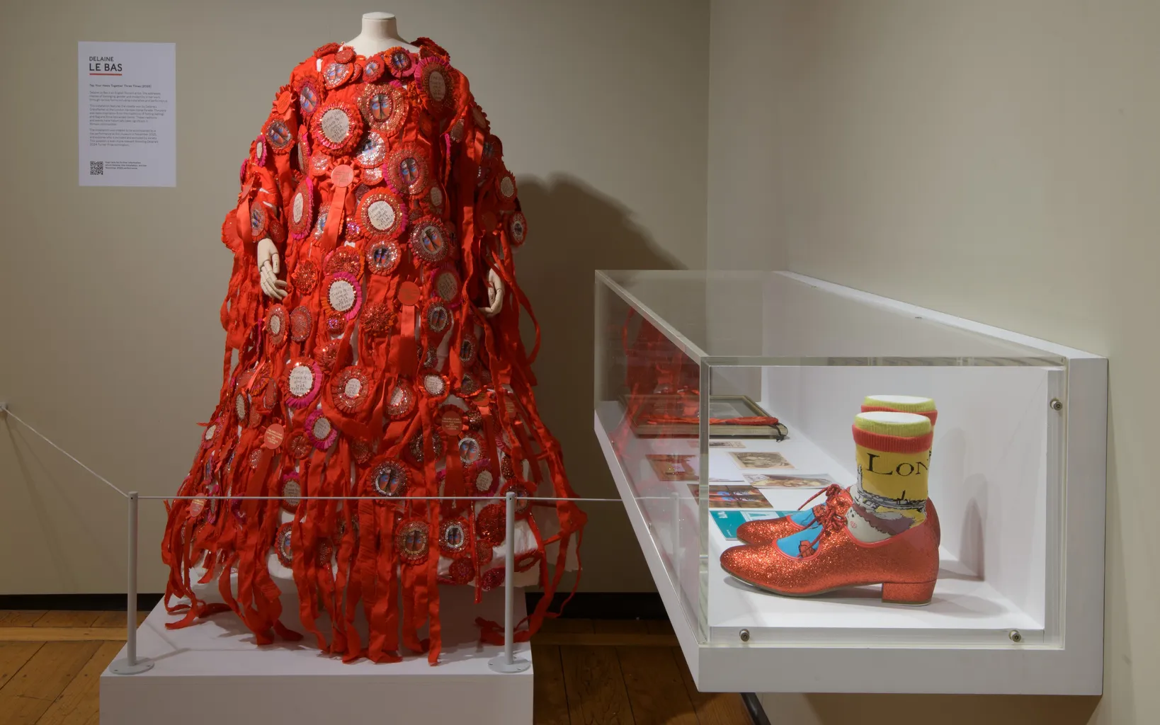 A mannequin displays a red, ribbon-covered dress with circular patches next to a glass case containing glittery red shoes and colorful socks.