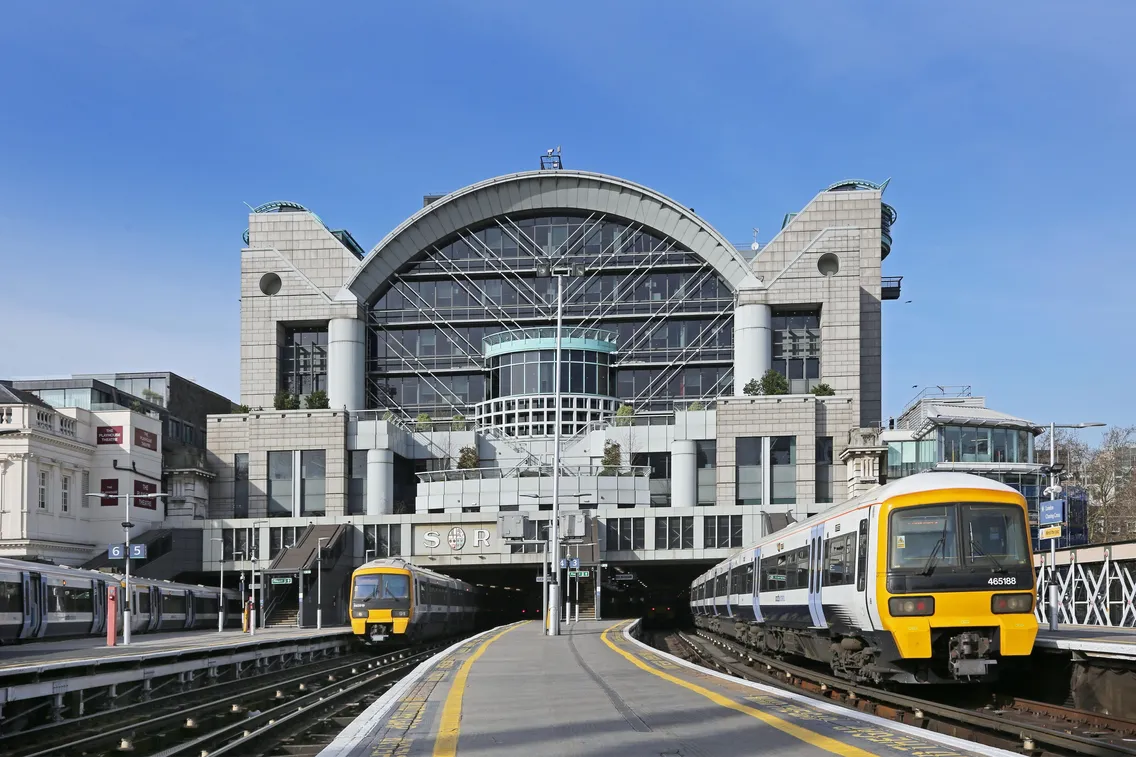 Two trains are at platforms outside a large, modern glass and steel railway station building with arched roof and "SOR" signage on the front.