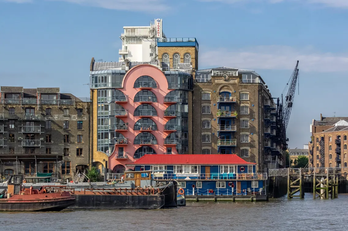 Historic waterfront buildings with a distinctive pink and white arched facade, docked boats, and an old crane along the Thames River under a blue sky.