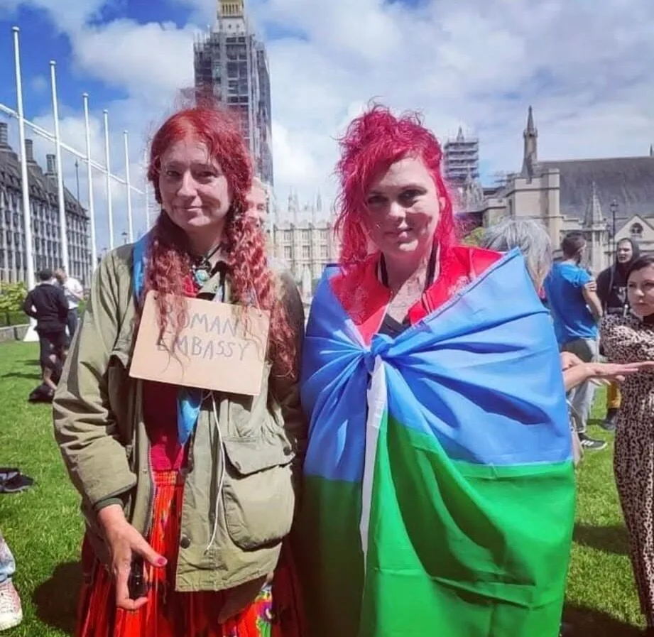 Two women with red hair stand on grass; one wears a cardboard sign reading "Romani Embassy," the other is draped in a blue and green flag. Buildings and people are in the background.