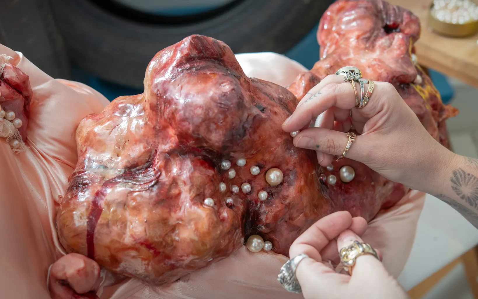 Hands attaching pearls to a large, fleshy, organic-looking sculpture with a pink base, possibly as part of an art installation or project.