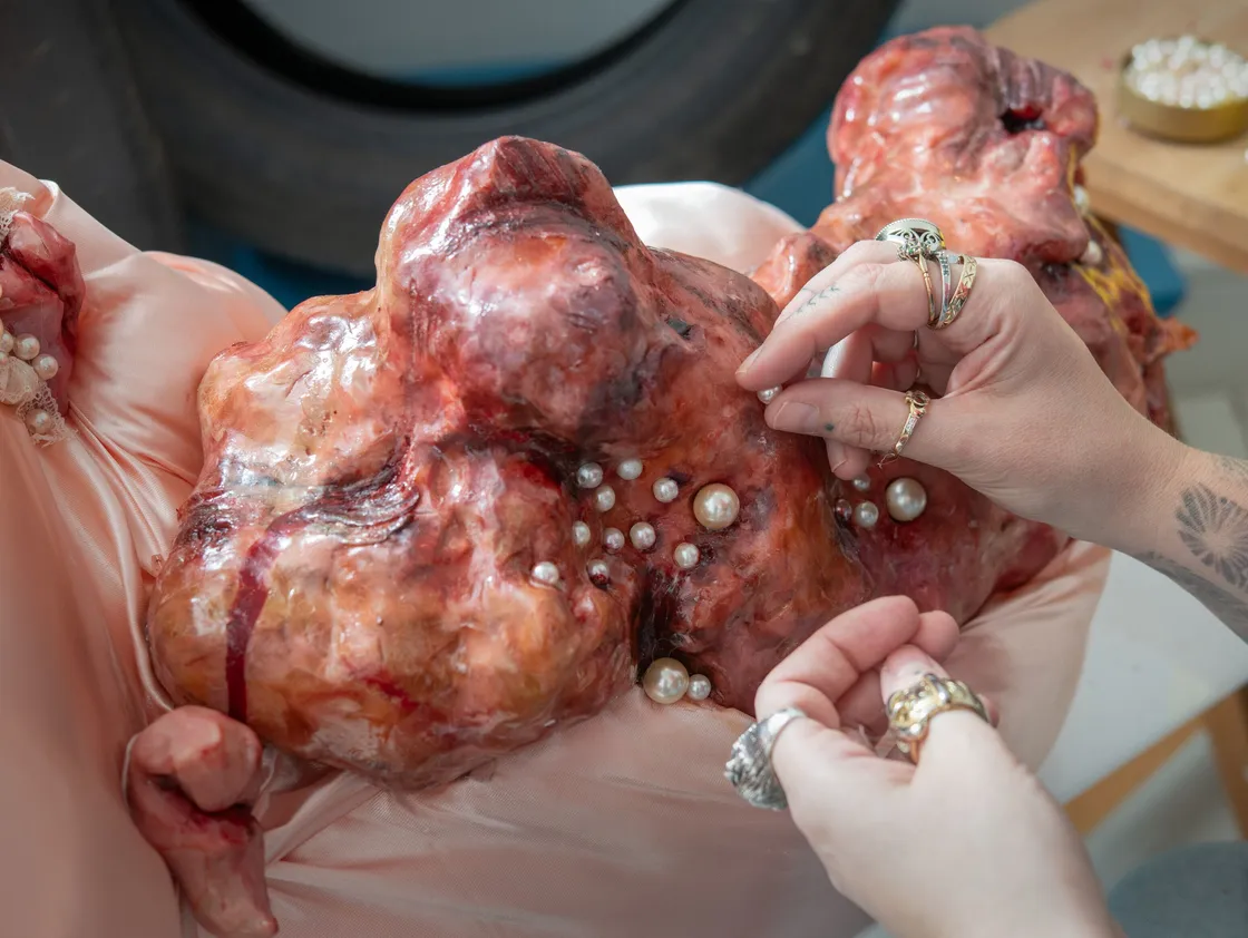 Hands attaching pearls to a large, fleshy, organic-looking sculpture with a pink base, possibly as part of an art installation or project.