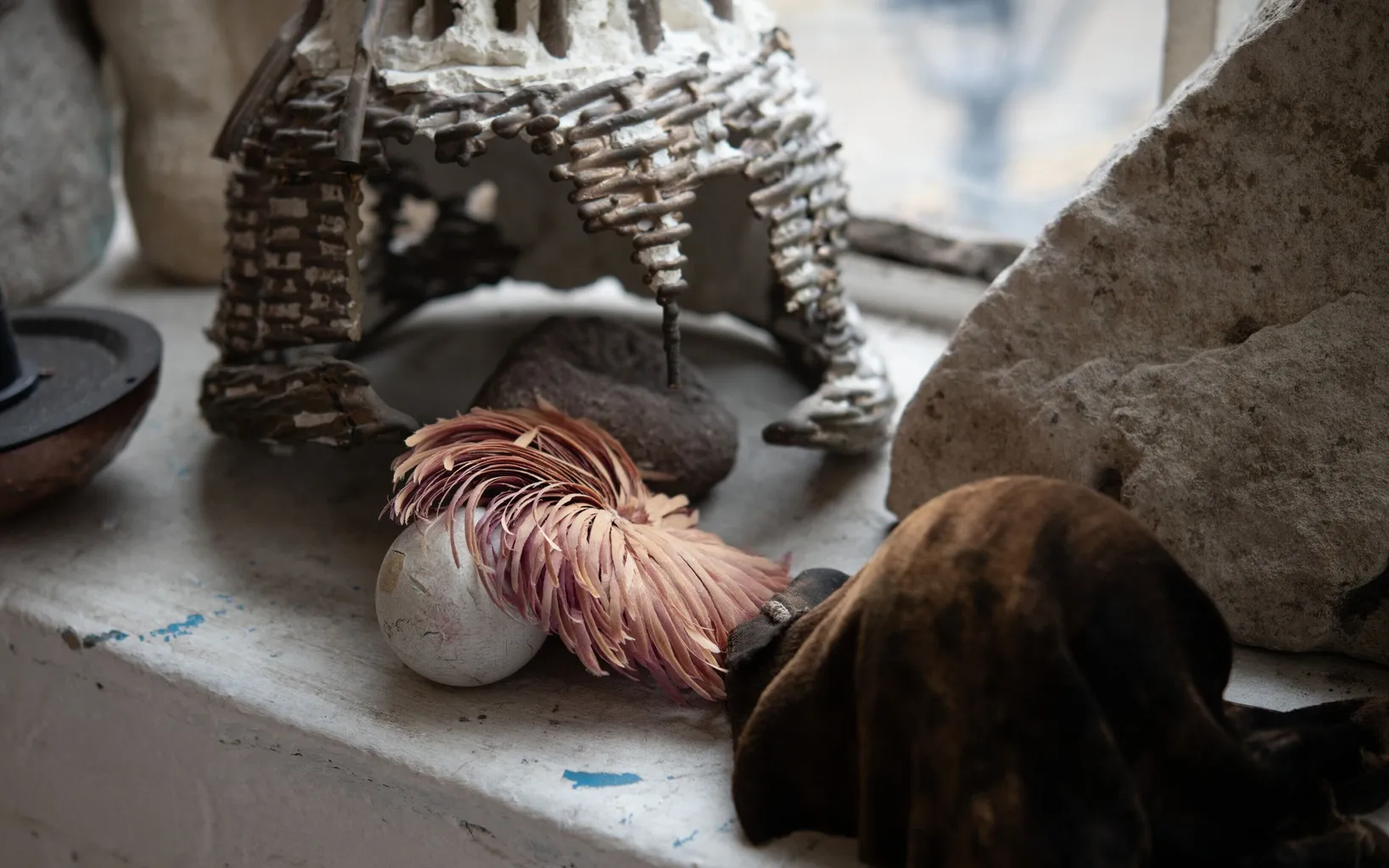 A white and pink feathered object, a white sphere, rocks, and a small model structure are arranged on a windowsill.
