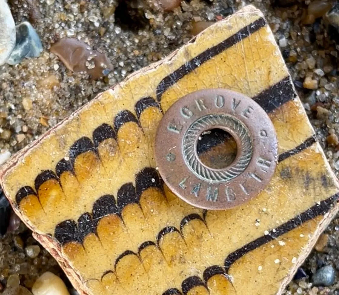 A round metal washer stamped with “GROVE OF LAMBETH” rests on a patterned ceramic fragment amid small pebbles and sand.