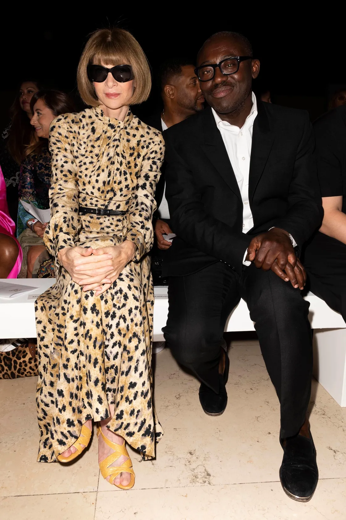 A person in a leopard print dress and sunglasses sits next to another in a black suit on a white bench.