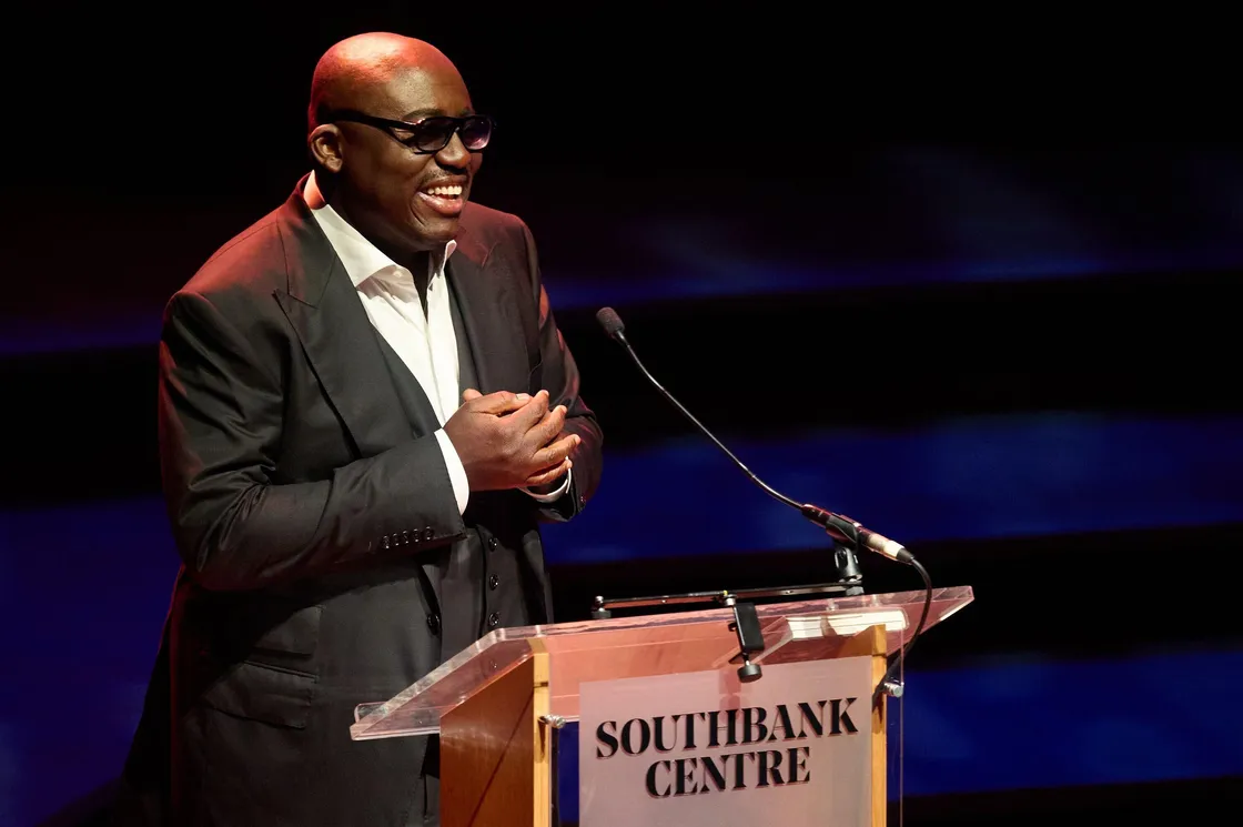 A man in a dark suit and sunglasses speaks at a podium labeled "Southbank Centre" on a stage with dim lighting.