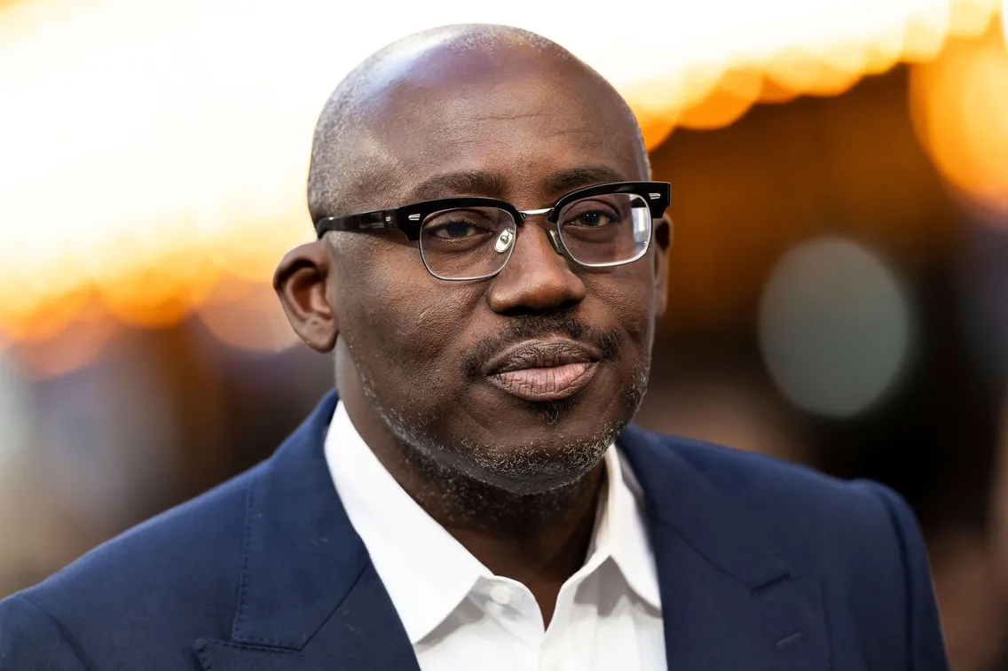 A person wearing glasses and a suit is looking at the camera with blurred lights in the background.