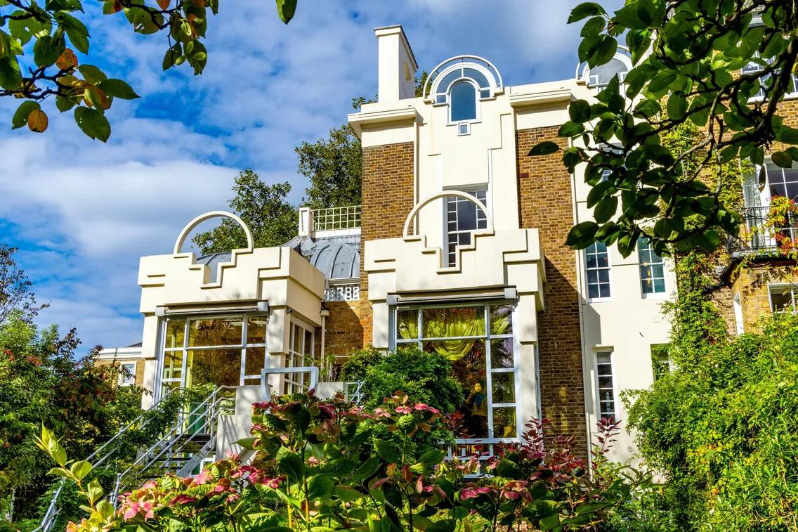 A modern house with large windows, geometric architectural details, and surrounded by green foliage and flowering plants under a partly cloudy sky.