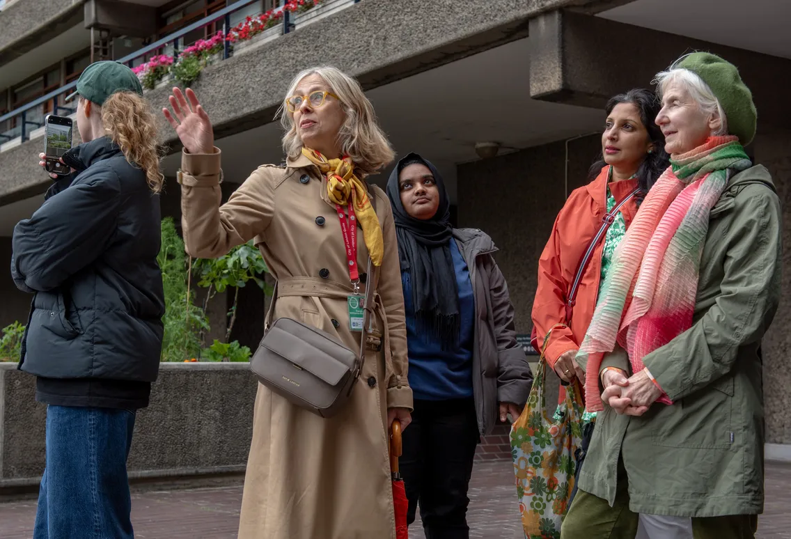 A group of five women stand outdoors near a concrete building; one gestures while speaking, others listen, and one holds a camera.