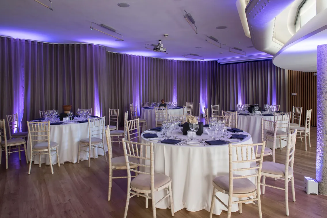 A banquet hall with circular tables set for a formal event. Each table has white tablecloths, wine glasses, and napkins. The room is lit with purple accent lighting and features wooden floors.