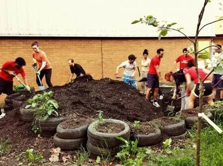 Group of people working together, shoveling dirt from a large pile into wheelbarrows, with stacked tires in the foreground and a brick building in the background.