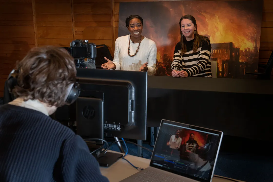 A man and a woman are seated and speaking; the woman gestures with her hands. Behind them is a backdrop depicting a historical fire scene. A tablet is placed on the table in front of the man.