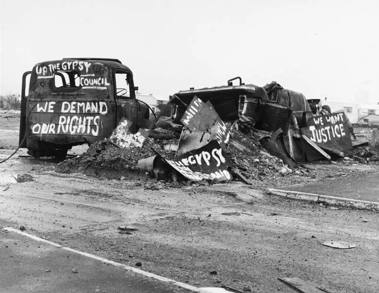 Black and white photo of a demolished vehicle with protest signs saying "we demand our rights" and "we want justice" at a barren site.