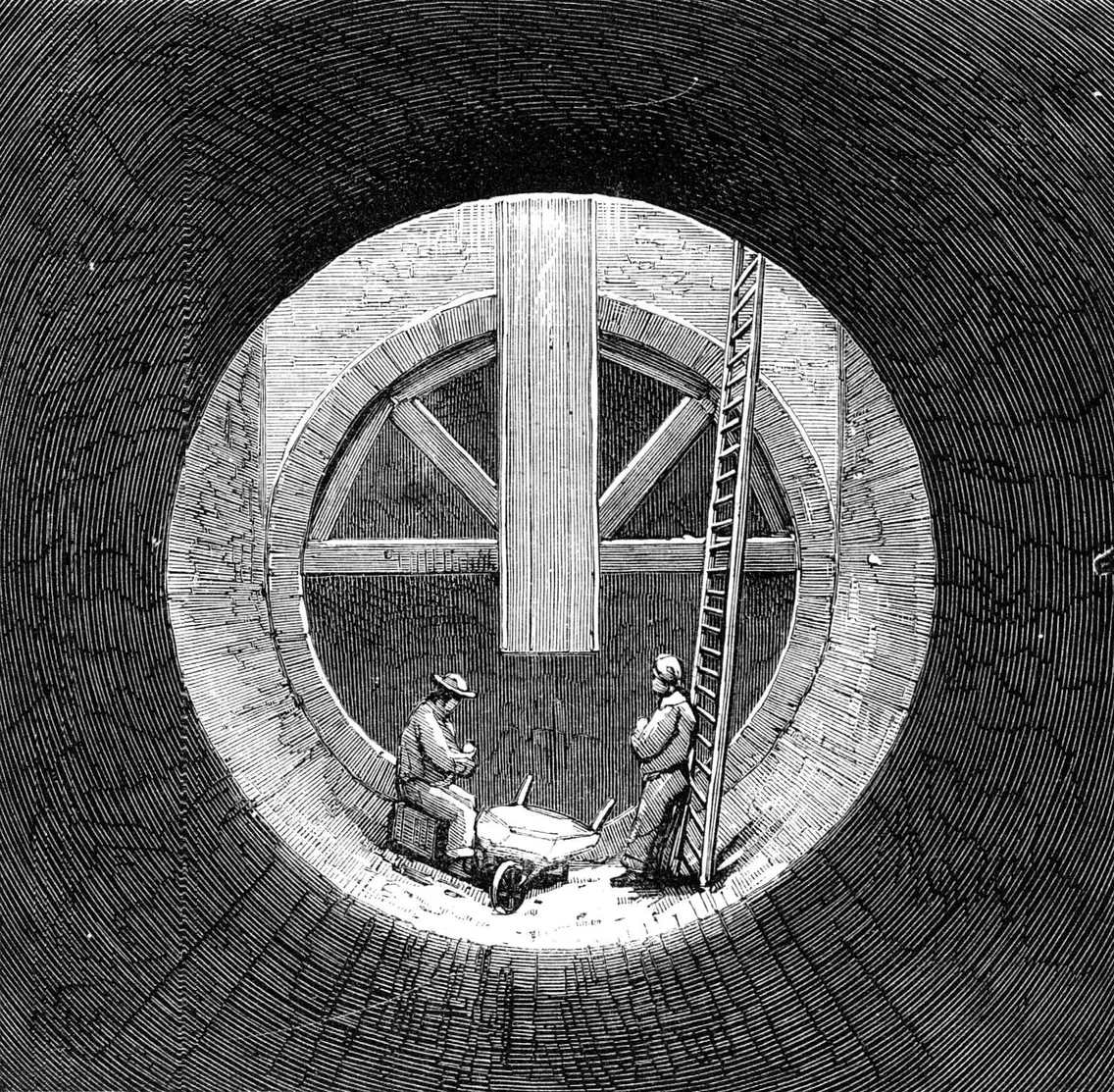 Two workers stand at the bottom of a large, circular shaft with a ladder and a wheelbarrow; viewed from inside the shaft, with brickwork and beams visible.
