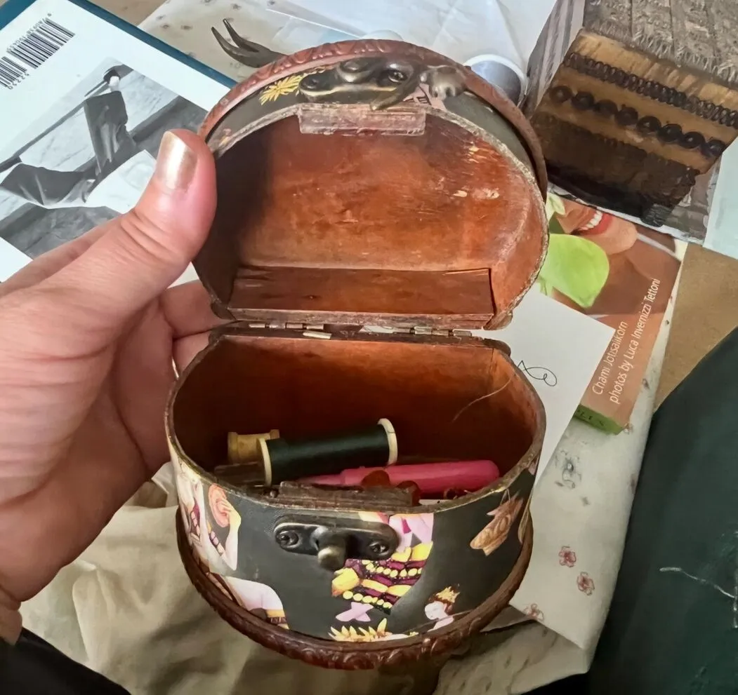 A hand holds an open small, decorated wooden box containing two spools of thread and a thimble.