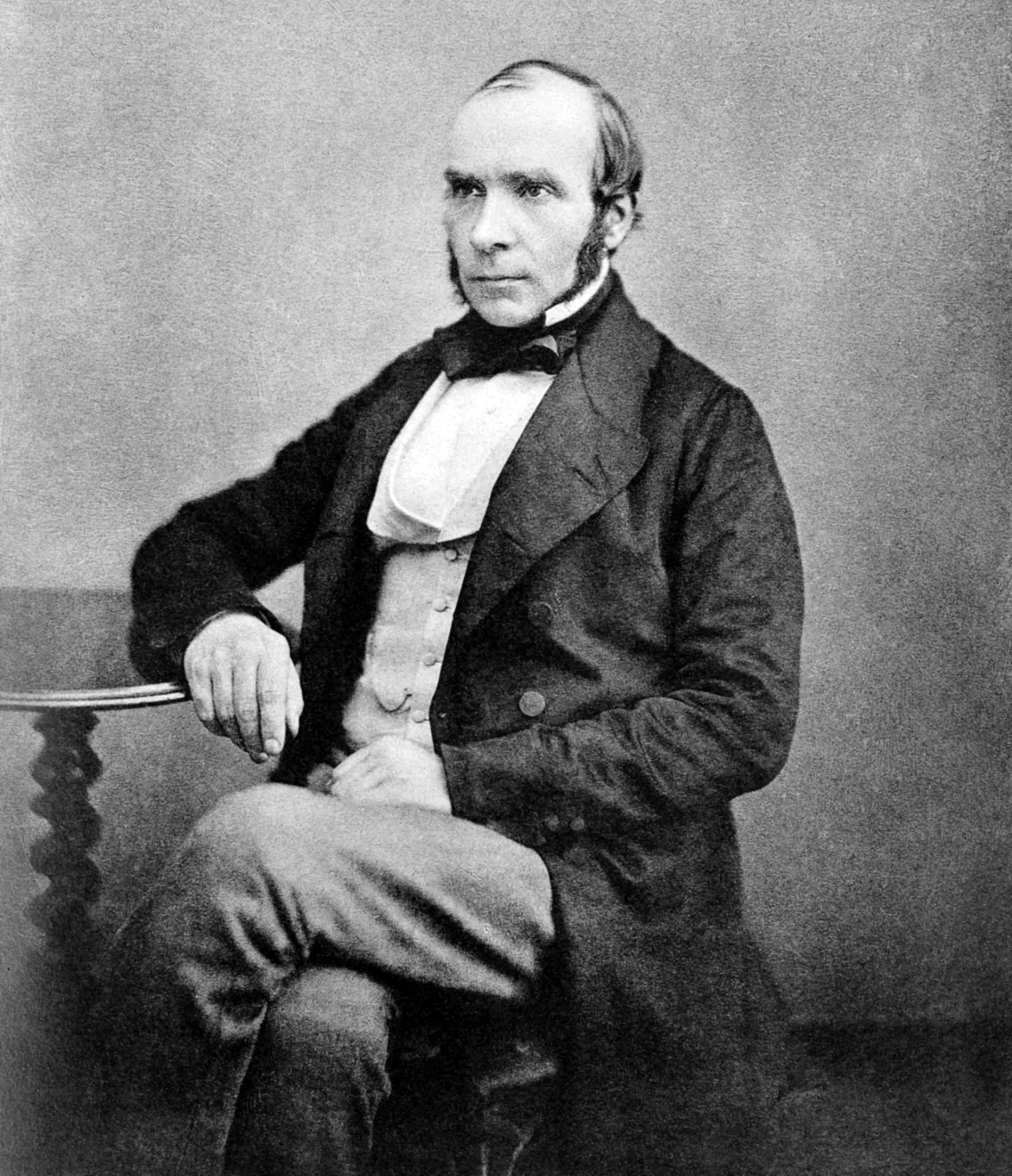 Black and white portrait of a seated man in 19th-century attire, looking slightly to the side with one arm resting on a table.