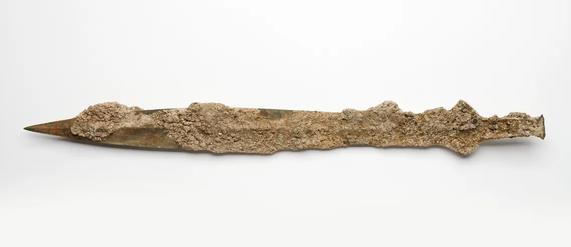 An ancient, corroded short sword with a pointed end and textured surface, lying horizontally against a plain white background.