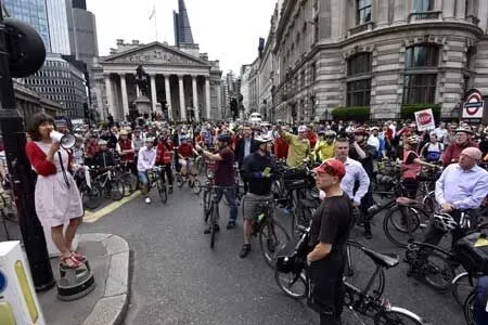 A woman with a megaphone addresses a crowd of cyclists gathered on a city street near historical buildings.