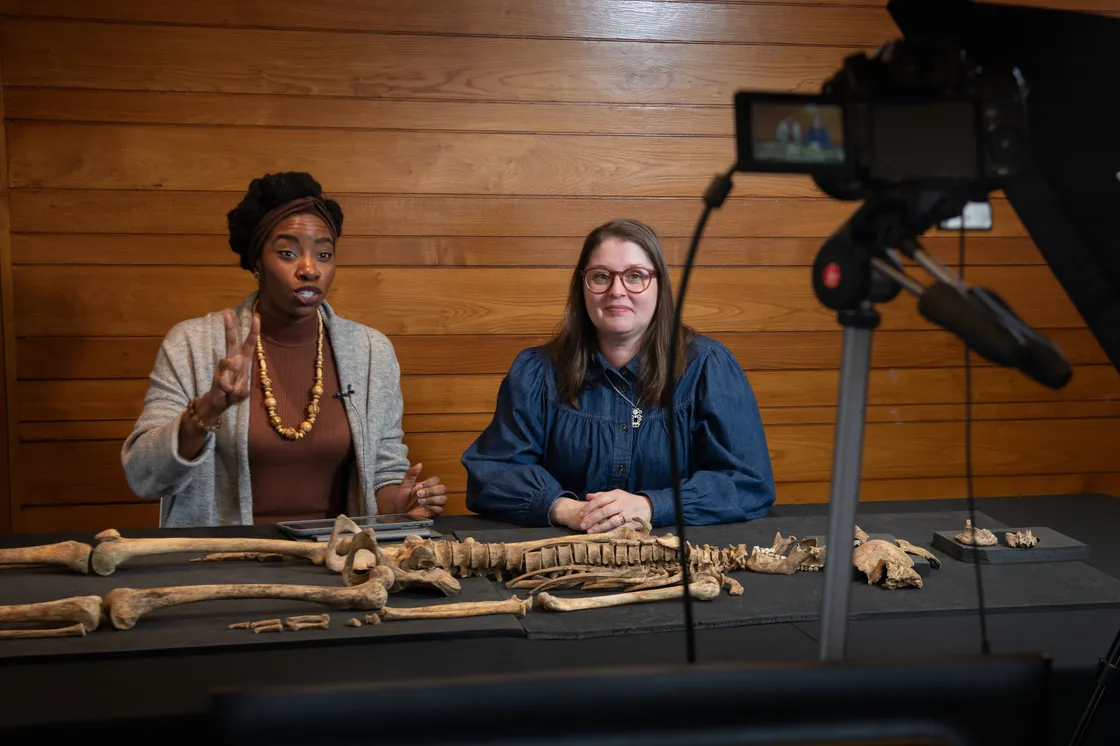 Two people seated at a table examining human skeletal remains, with a full skeleton model and a decorative backdrop in the background.