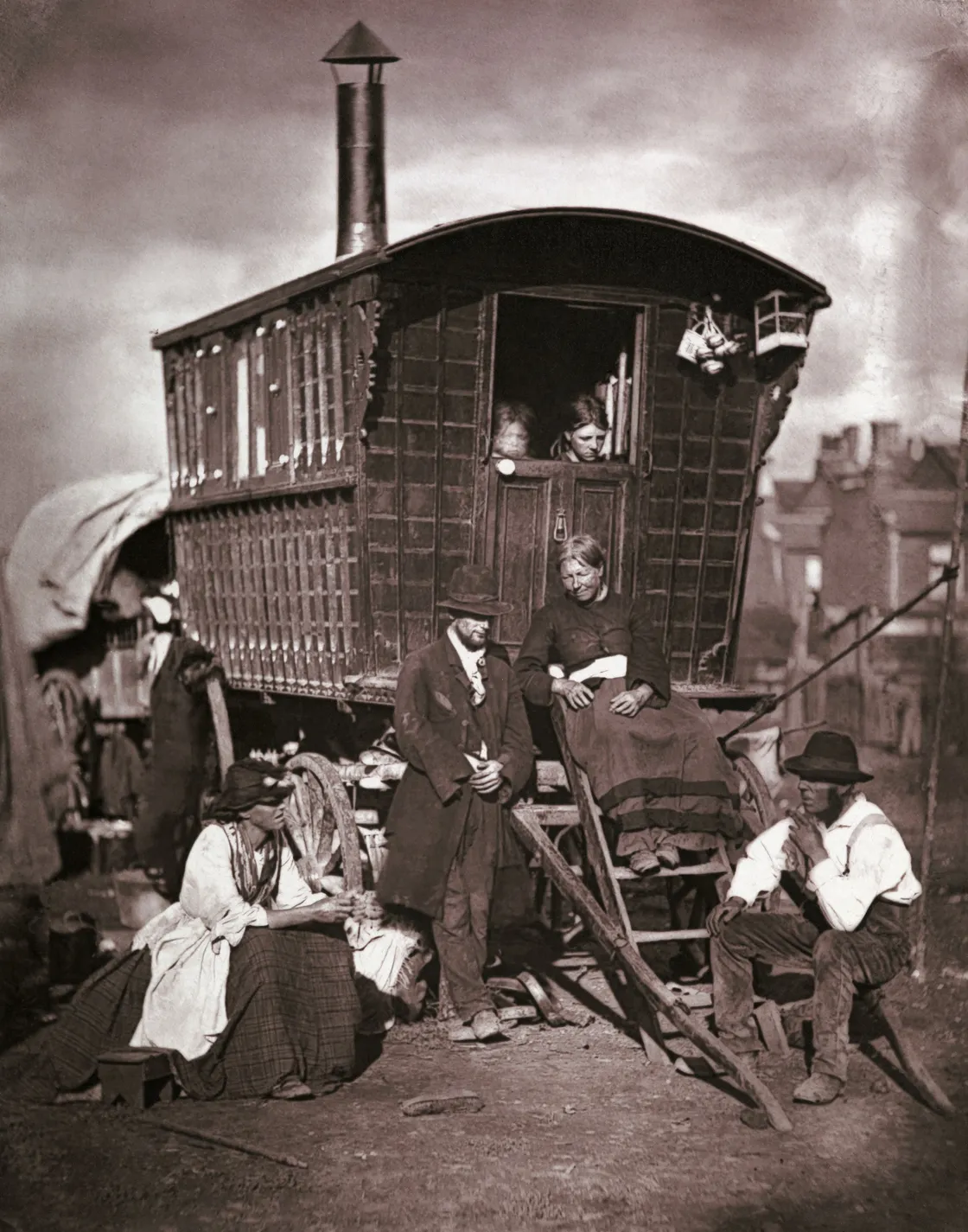 A vintage photo of a family with children peeking from a wooden caravan, surrounded by adults in period attire siting and standing outside.