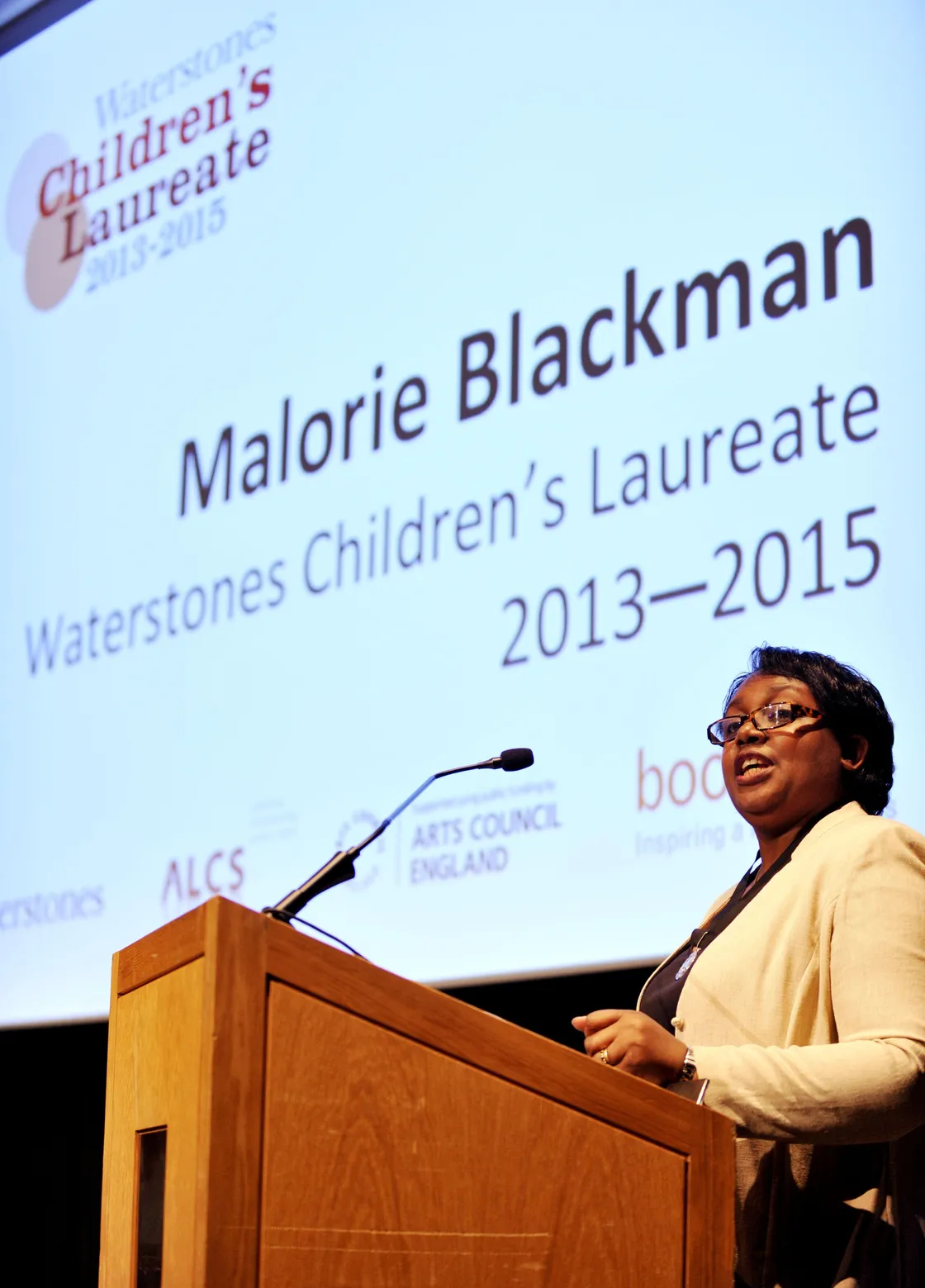 A person stands at a podium with a projector screen displaying "Malorie Blackman, Waterstones Children's Laureate 2013-2015" in the background.