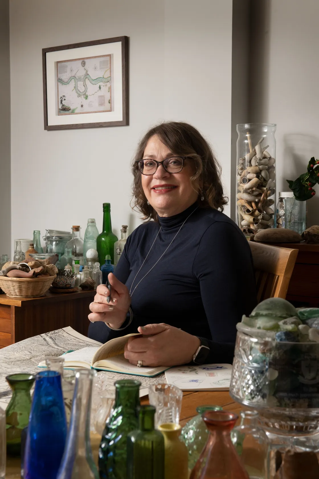 A woman with glasses sits at a table writing in a notebook, surrounded by colorful glass bottles and shells, with framed art on the wall behind her.