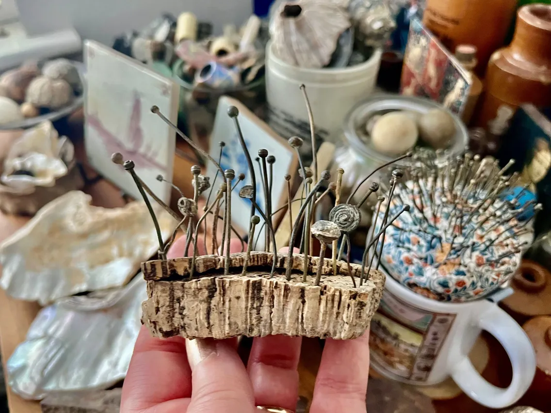 A hand holds a piece of cork with small nails and tiny snail shells attached; various shells and objects are scattered on a cluttered surface in the background.
