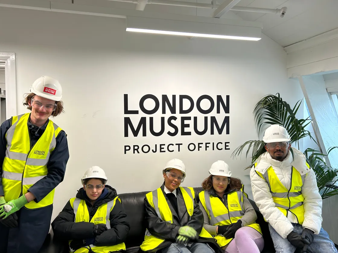 Five young people in high-vis yellow jackets and hard hats sitting in front of a sign that reads 'London Museum project office'