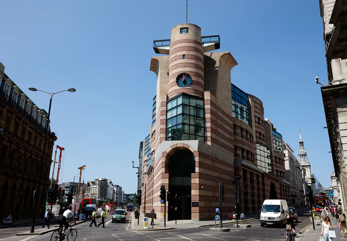 A modern, striped office building with a central tower and rounded corners stands at a busy city intersection under a clear blue sky. People, cars, and cyclists move along the street.