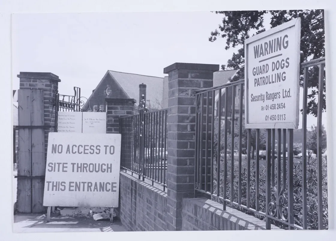 Brick gate entrance with signs reading "NO ACCESS TO SITE THROUGH THIS ENTRANCE" and "WARNING: GUARD DOGS PATROLLING" by Security Rangers Ltd. A brick building and tree are visible in the background.